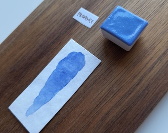 Può includere: Un set di acquerelli con un blocco di vernice quadrato blu e un campione di vernice blu su carta bianca. La parola "PROVENCE" è su una piccola etichetta bianca. Gli elementi sono su una superficie di legno.