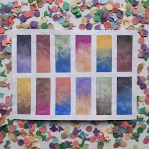 May include: A watercolor paint swatch card with 12 different color swatches. The swatches are arranged in a grid of four rows and three columns. The colors are a mix of blues, purples, pinks, and greens.