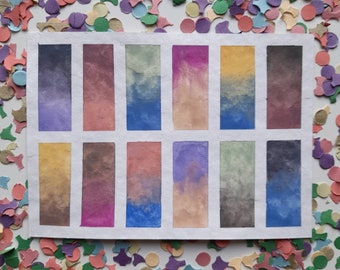May include: A watercolor paint swatch card with 12 different color swatches. The swatches are arranged in a grid of four rows and three columns. The colors are a mix of blues, purples, pinks, and greens.
