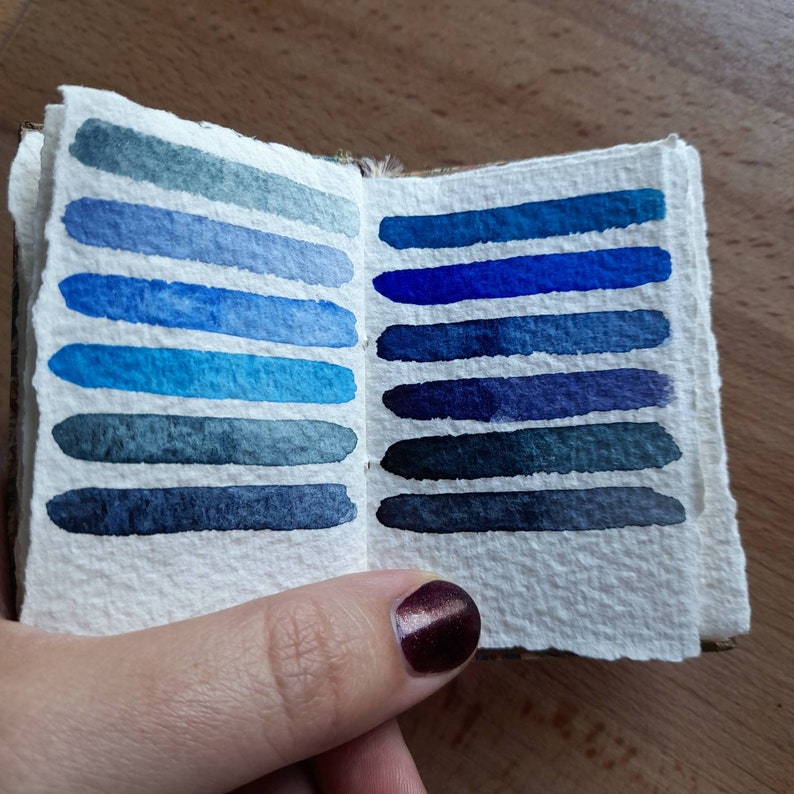 May include: A small, open book with watercolor swatches of blue and grey shades. The book is made of textured paper and has a brown cover.