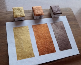 May include: Three watercolor paint pans with metallic pigments. The pans are labeled "Pure Gold", "Burnt Orange", and "Bronze". The paint swatches are shown below the pans.