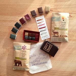 May include: A set of watercolor paints in a variety of colors, including red, brown, green, and gray. The paints are arranged in a circle on a wooden surface. There are also several small bags with labels that say "Clematis" and "Calla Lily".