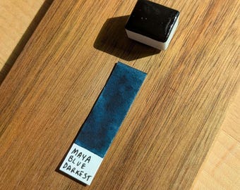 Può includere: Un cubo di pittura ad acquerello nero con base bianca è posizionato accanto a un campione blu su una superficie di legno. Il campione riporta la scritta "MAYA BLUE DARKEST". La venatura del legno è visibile.
