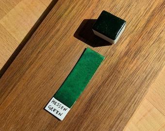 Peut inclure: Une planche de bois avec un échantillon de peinture aquarelle vert foncé étiqueté "HESSEN GREEN" et un petit bloc de peinture carré. L'échantillon est vert vif, et le bloc est d'un vert plus foncé.