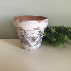 May include: White terracotta planter with a distressed finish and a black and white bee design. The planter is decorated with a wreath of leaves and a bee in the centre.