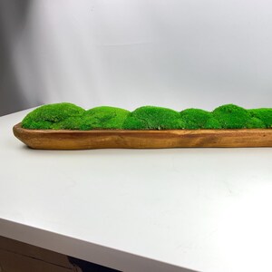 May include: A long, wooden trough planter filled with artificial green moss. The planter is made of natural wood and has a rustic, farmhouse style.