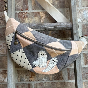 May include: A crescent-shaped crossbody bag featuring a patchwork design of dark gray denim, light peach fabric, and cream fabric with black polka dots. The bag includes two white ghost appliqués and a metal zipper pull shaped like a pumpkin.