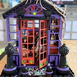 Peut inclure: Une structure de maison miniature violette et orange avec des accents de papillons noirs, deux portes orange ouvertes avec des autocollants de chats noirs, des ornements en gemmes vertes et des fleurons noirs sur la base avant avec un tapis.