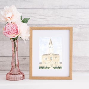 Op de afbeelding: Een ingelijste aquarelprint van een gebouw met een hoge torenspits, tegen een lichtblauwe lucht. Het kunstwerk zit in een lichte houten lijst. Links van de lijst staat een roze glazen vaas met roze pioenrozen.