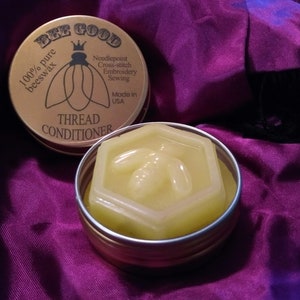 May include: A round tin container with a gold lid and a label that reads "Bee Good 100% pure beeswax Thread Conditioner". The label also includes the text "Needlepoint Cross-stitch Embroidery Sewing Made in USA". The tin contains a hexagonal block of yellow beeswax.