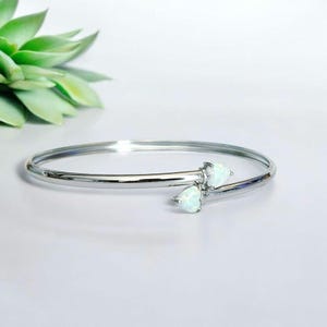 Puede incluir: Un brazalete plateado con dos piedras de ópalo blanco en forma de pera. El brazalete tiene un diseño simple y elegante y se exhibe sobre una superficie blanca. La joyería es un accesorio elegante.