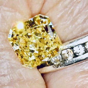 May include: A close-up of a yellow diamond ring. The large, rectangular-cut diamond is the focal point, with a brilliant, faceted surface. The ring's band is silver, adorned with smaller, clear diamonds.