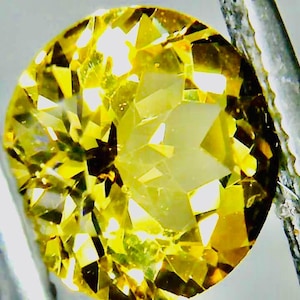May include: A faceted, round-cut yellow gemstone, held by metal tweezers. The gem has a brilliant, multi-faceted surface that reflects light, showcasing a range of yellow hues. The close-up shot highlights the gem's clarity and intricate cut.