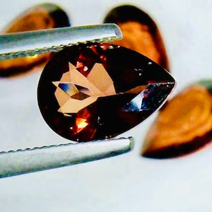 May include: A faceted, teardrop-shaped gemstone held by tweezers. The gem is a deep brown color with lighter brown and reflective facets. Two other gems are visible in the background, along with a white surface.