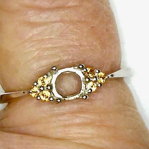 May include: A silver ring with a gold-colored floral design on the setting. The ring features a light brown, oval-shaped stone. The ring is on a finger.