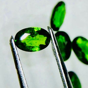 May include: A close-up of a green oval gemstone held by tweezers. The gemstone is faceted and has a bright, vibrant green color.