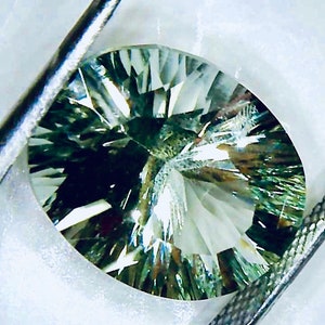 May include: A green and white gemstone with a starburst pattern. The gemstone is oval shaped and has a high polish.
