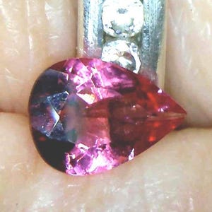 May include: A pear-shaped, faceted gemstone with a deep magenta and reddish-pink hue. The stone is set in a metallic band featuring two small, round, clear white accent stones positioned vertically above the main pear-shaped gem.