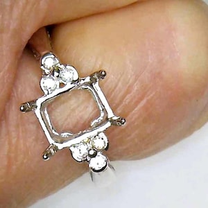 May include: Close-up of a silver ring setting with a square center and small, round accent stones. The ring setting is held by a person's hand, showcasing the intricate design and potential for a custom gemstone.