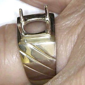 May include: A gold-toned ring setting with an oval-shaped space for a gemstone. The ring has a rectangular top with four prongs and a textured band with diagonal lines. The ring is held by a person.