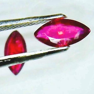 May include: Two marquise-cut, vibrant red gemstones. One is held by silver tweezers, showcasing its polished surface and rich color. The other gem is positioned nearby, also displaying its deep red hue against a white background. These gems are ideal for jewelry making.