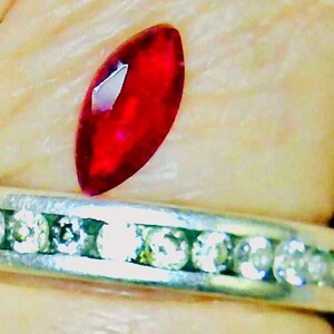 May include: A marquise-cut, deep red gemstone rests above a silver band ring with a row of small, clear gemstones. The red gemstone has a reflective surface. The ring is set against a neutral background.