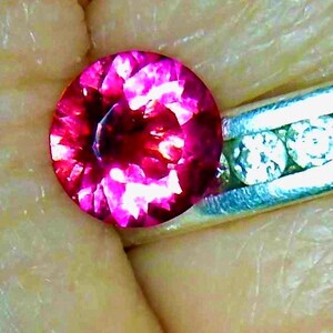 May include: A round, faceted, deep pink gemstone, possibly a ruby or tourmaline, is set next to a silver ring with small, clear stones. The gemstone is held in a hand, with the skin tone visible in the background.