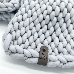 May include: A close-up shot of a light grey, chunky knit item. The thick, braided texture is prominent, giving a soft, tactile feel. A small, dark grey tag with the text "BIRCH & CIDER" is attached. The item appears to be a home decor item.