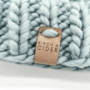 Peut inclure: Un bonnet en tricot bleu clair avec une étiquette en cuir marron qui dit "Birch & Cider".