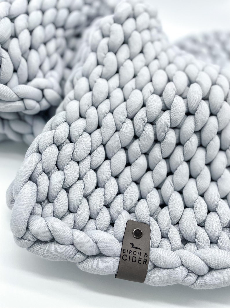 May include: A close-up shot of a light grey, chunky knit item. The thick, braided texture is prominent, giving a soft, tactile feel. A small, dark grey tag with the text "BIRCH & CIDER" is attached. The item appears to be a home decor item.