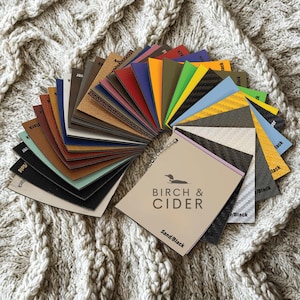 May include: A selection of material samples in various colours, including black, blue, red, and yellow, with a Birch & Cider tag. The samples are displayed on a textured, off-white surface.