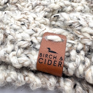 Peut inclure: Gros plan sur un textile tricoté épais de couleur crème, beige et noir. Une étiquette en cuir marron est attachée, avec le texte "BIRCH & CIDER" et une silhouette de chien. Le tissu tricoté semble doux et texturé, pour une écharpe ou une couverture.