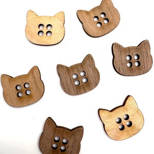 May include: A collection of wooden cat-shaped buttons in various shades of brown. Each button features four evenly spaced holes for sewing. The buttons vary in color from light tan to dark brown, with visible wood grain.