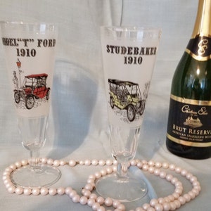 May include: Two vintage glass champagne flutes with illustrations of a Model T Ford and a Studebaker automobile from 1910. A bottle of Chateau Ste. Michelle Brut Reserve American Sparkling Wine is in the background.