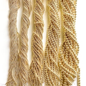 May include: Five strands of gold-colored beads, each with a different texture and size. The beads are arranged vertically and are all the same color.