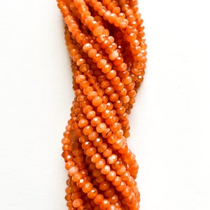 May include: Strand of faceted orange gemstone beads. The beads are small, round, and have a sparkling, polished appearance. The beads are strung together, creating a flowing, textured effect. The background is plain white.