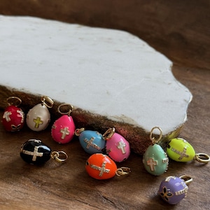 May include: A collection of ten small, colorful egg-shaped charms with gold accents and a cross design. Each charm has a different color, including white, pink, black, blue, orange, green, and purple.
