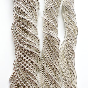 May include: Three strands of silver beads, each strand is made up of small, round beads. The beads are arranged in a loose, flowing pattern.