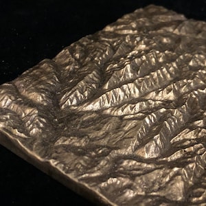 May include: A detailed, metallic relief sculpture of a mountain range. The piece is rectangular, with a textured surface depicting peaks, valleys, and ridges. The color is a muted silver-bronze, and the lighting creates shadows that emphasize the topography.