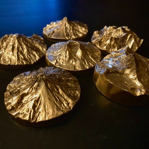 May include: Six metallic gold-colored coasters, each shaped like a mountain range. The coasters have a textured surface, with peaks and valleys. The coasters are arranged in a circular pattern on a dark surface.