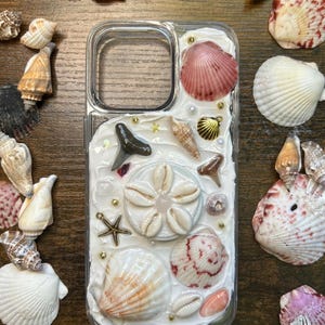 May include: Clear phone case decorated with various seashells, including pink, white, and tan varieties. The case features a white textured background with gold accents and a small shark tooth. Seashells are also scattered around the case.