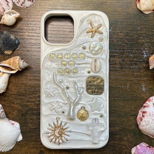 Może przedstawiać: Białe etui na telefon z teksturowanym wzorem przedstawiającym rozgwiazdę, muszle i słowa "HOLD GOLD GOD". Etui ma złoty krzyż, gałązkę koralowca i małą metalową plakietkę z napisem "DREAMING OF THE SEA".