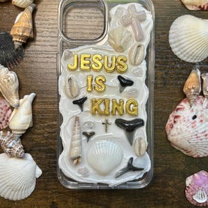 Może przedstawiać: Przezroczyste etui na telefon ozdobione muszlami, krzyżami i złotym napisem „JESUS IS KING”. Etui otoczone jest różnymi muszlami na ciemnej drewnianej powierzchni.