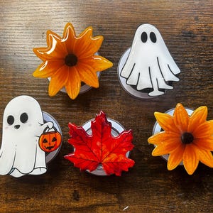 May include: Five circular phone grips featuring seasonal designs: two orange flowers with dark centers, two white ghosts with black features, one of which carries a small orange jack-o'-lantern bucket, and one vibrant red maple leaf.