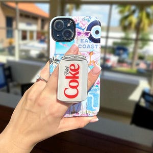 May include: A smartphone case featuring a collage of coastal-themed illustrations including palm trees, sunglasses, and an evil eye symbol, with the text East Coast. A circular grip attachment shaped like a silver Diet Coke can is centered on the back.