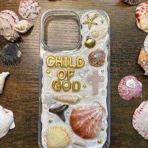 Może przedstawiać: Przezroczyste etui na telefon ozdobione muszlami, rozgwiazdą i złotym napisem "CHILD OF GOD". Etui ma białe, teksturowane tło i jest otoczone różnymi muszlami.
