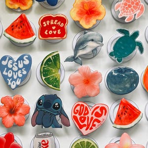 May include: A collection of circular phone grips featuring various designs including fruit slices like watermelon, lime, orange, and avocado, tropical flowers, sea turtles, a dolphin, a soda can, and hearts with the text Jesus Loves You and God Is Love.