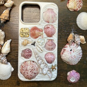 Może przedstawiać: Białe etui na telefon ozdobione różnymi muszlami w odcieniach różu, kremu i brązu. Etui ma teksturowany wzór i napis "Dreaming of the Sea". Rozgwiazda i inne muszle otaczają etui.
