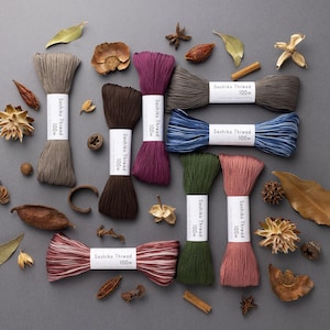 Puede incluir: Surtido de hilos Sashiko de 100 m en varios colores, incluyendo gris, marrón, burdeos, azul, verde y rosa. Los hilos están dispuestos sobre un fondo gris con elementos naturales como hojas, palitos de canela y anís estrellado.