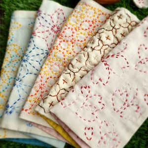 May include: A set of five folded fabric napkins featuring intricate sashiko-style embroidery in various colors including blue, yellow, orange, brown, and pink, arranged in a fan shape on a textured green surface.